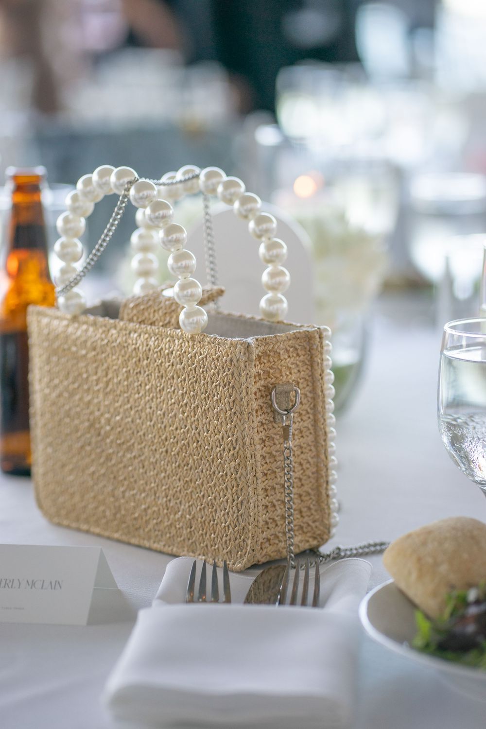 Woven straw handbag with pearl handles placed on a white linen wedding reception table.