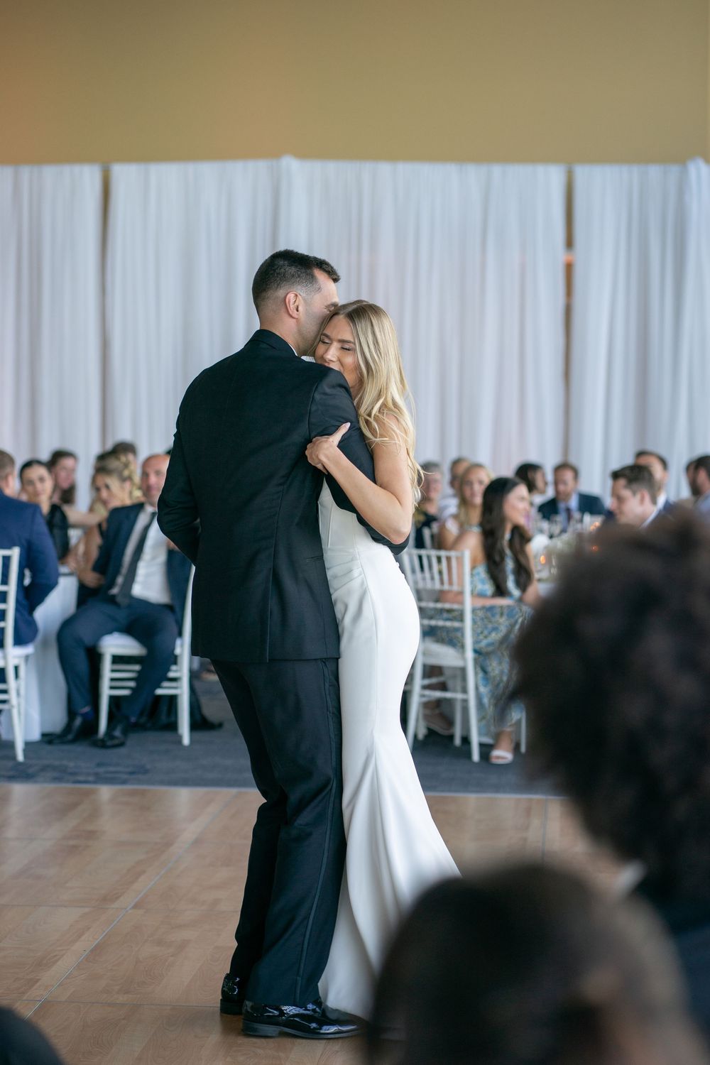 Newlyweds share an intimate first dance embrace while guests watch at elegant wedding reception.