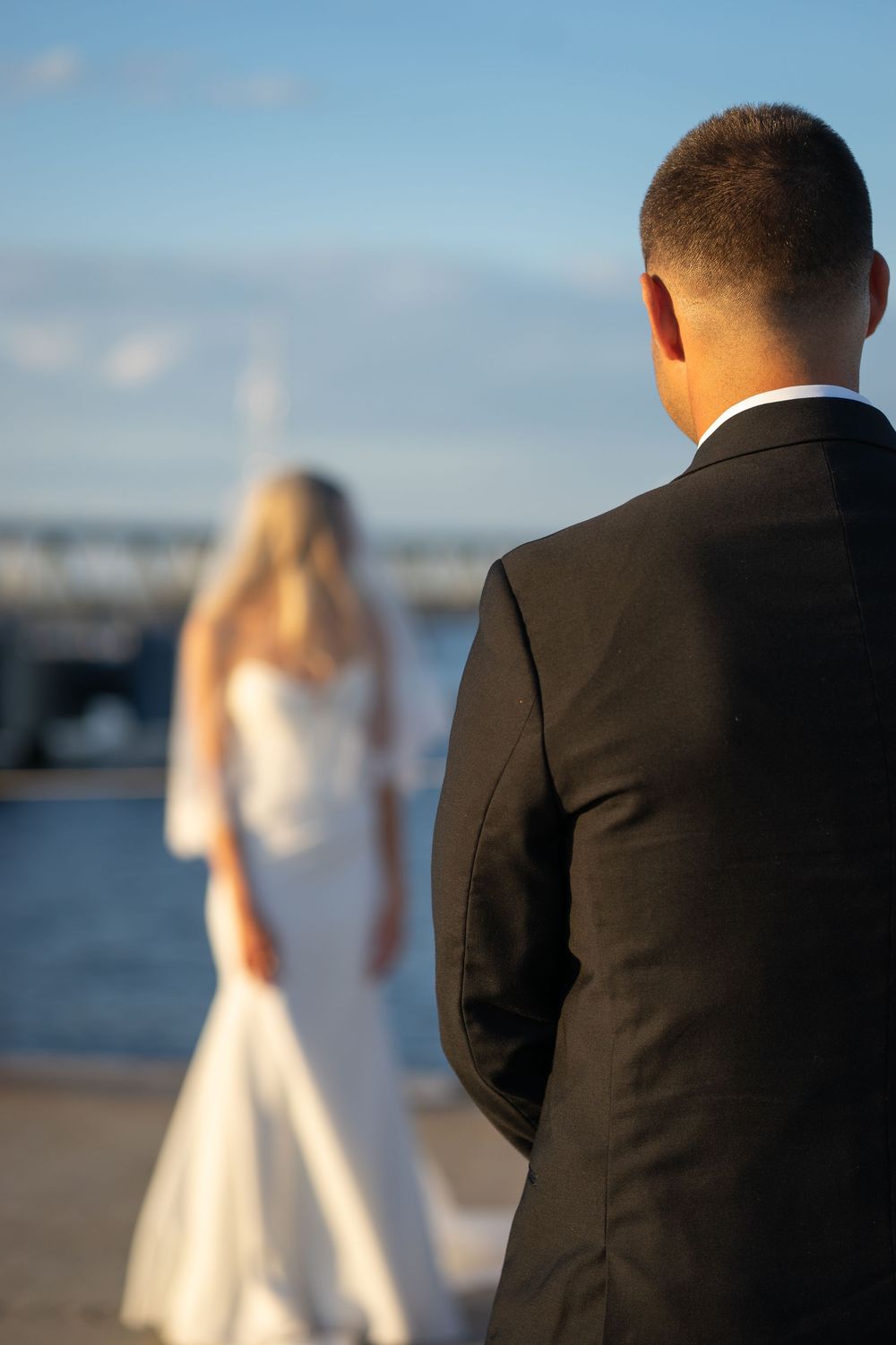 Groom in black suit watching his bride in a flowing white gown and veil on a waterfront dock.
