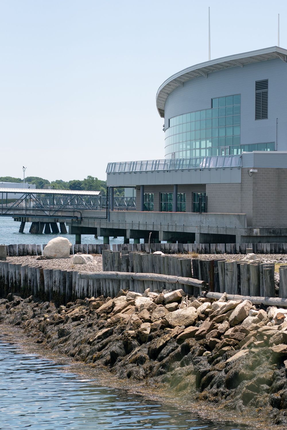 Modern waterfront event venue building on stilts with rocky shoreline and calm harbor waters.
