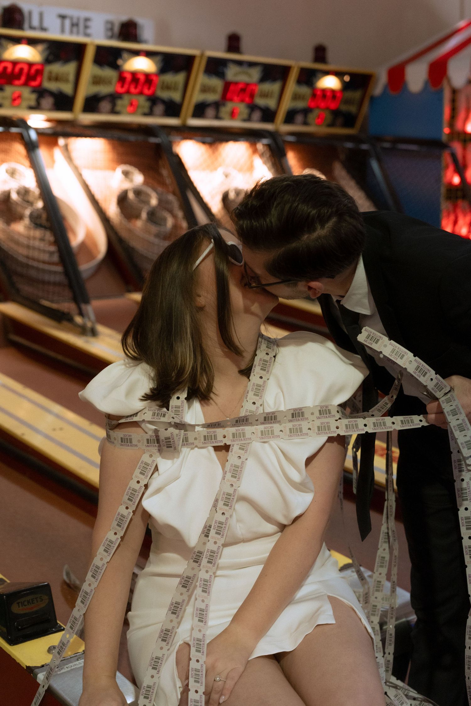 Couple in formal wear sharing a kiss surrounded by arcade tickets near skee-ball machines at a fun fair.