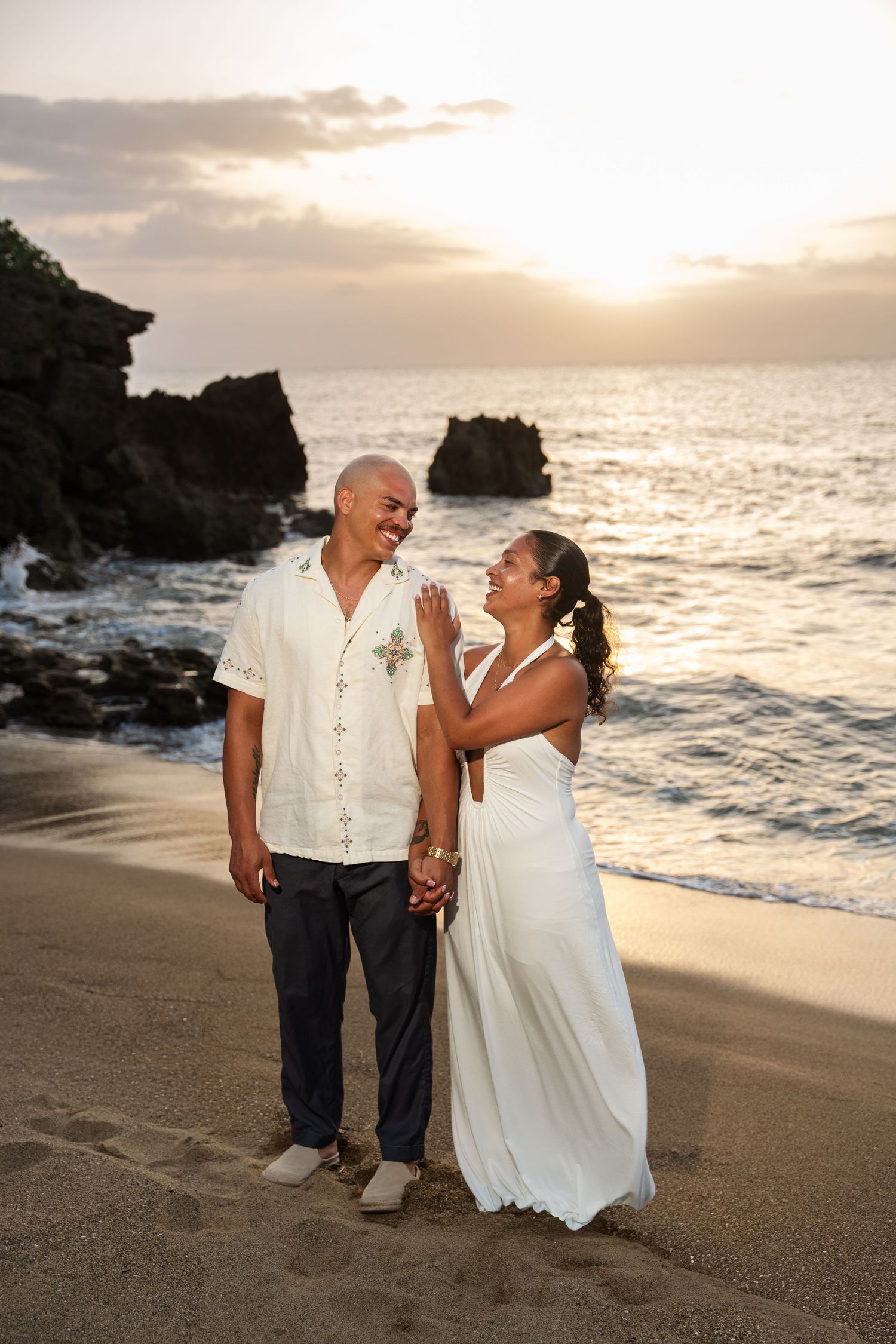 Couple in wedding attire sharing a romantic moment on a sandy beach at sunset with ocean waves behind them.