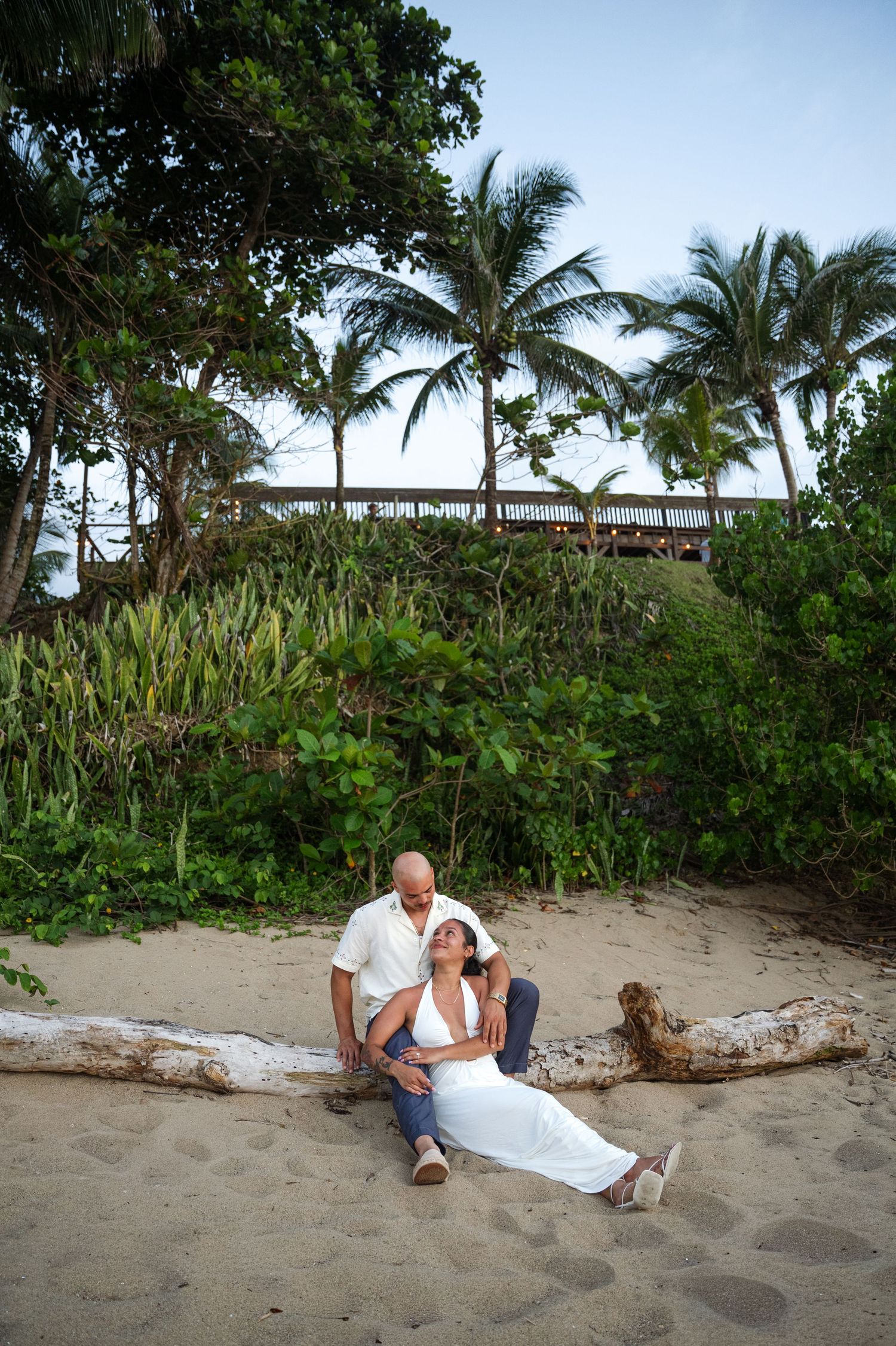 Romantic couple relaxing on sandy beach with palm trees, woman in white dress leaning on man near driftwood.