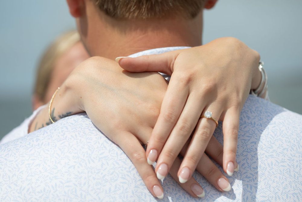 Close-up of a woman's hand with an engagement ring wrapped around a man's shoulder in a romantic embrace.