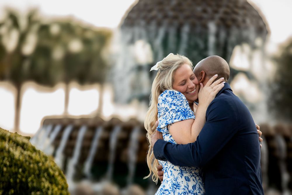 Couple embracing joyfully in front of a fountain, woman in blue floral dress and man in navy suit, outdoor engagement photo.