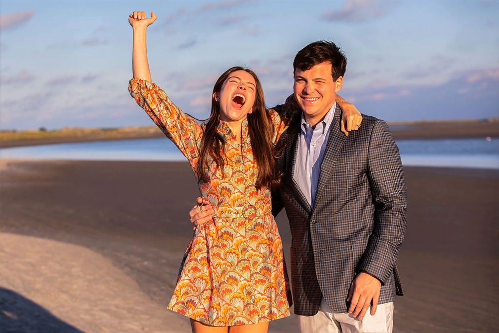 Joyful couple celebrating on a beach at sunset, woman raising fist in excitement, man in blazer smiling.
