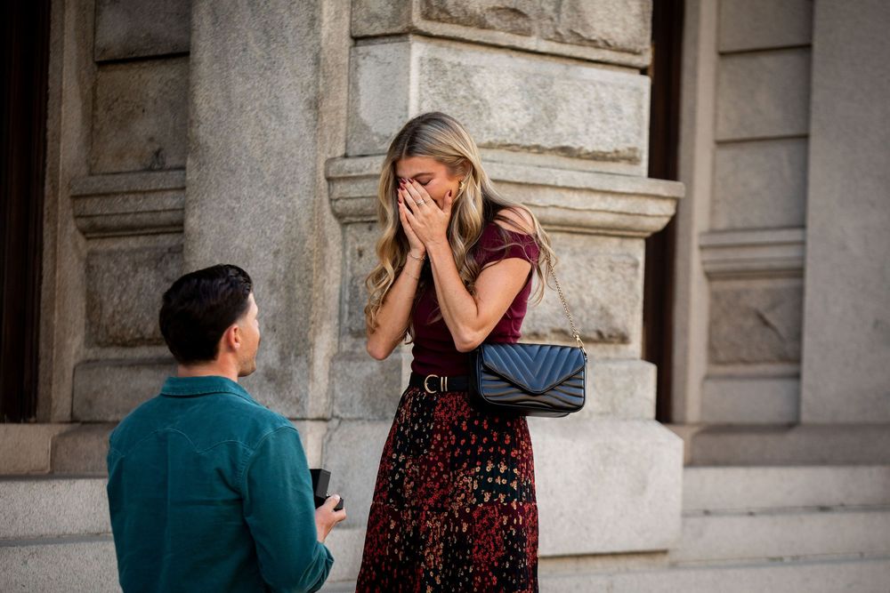 Man proposing to surprised woman in floral dress outside historic building, romantic marriage proposal moment.