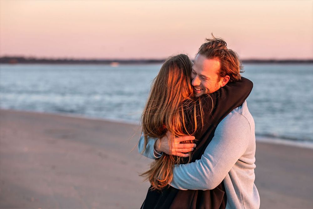 Couple embracing on a beach at sunset, smiling warmly with ocean waves and pink sky in the background.