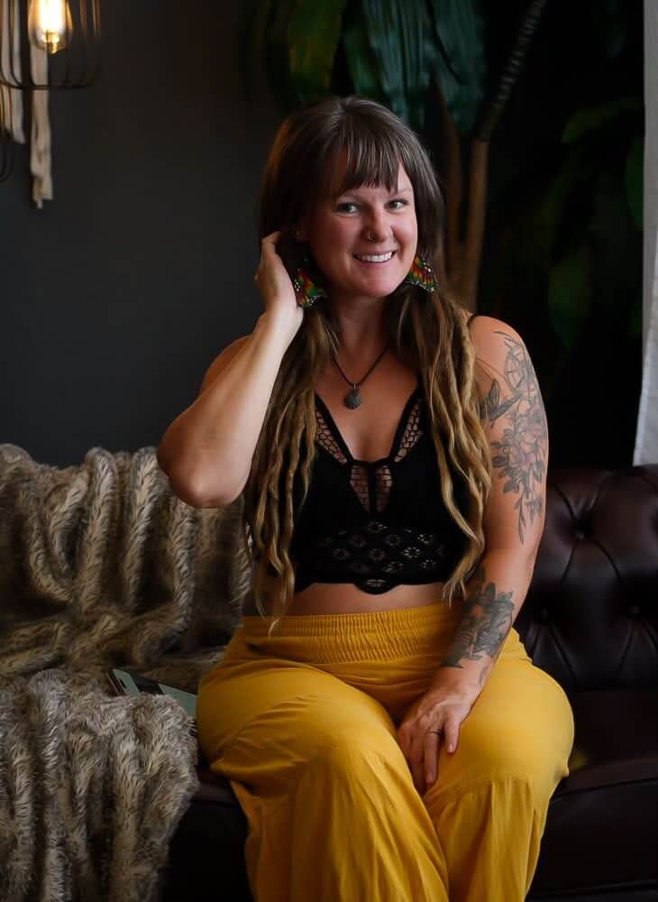 Smiling woman with dreadlocks, tattoos, wearing black crop top and yellow pants, sitting on leather couch.