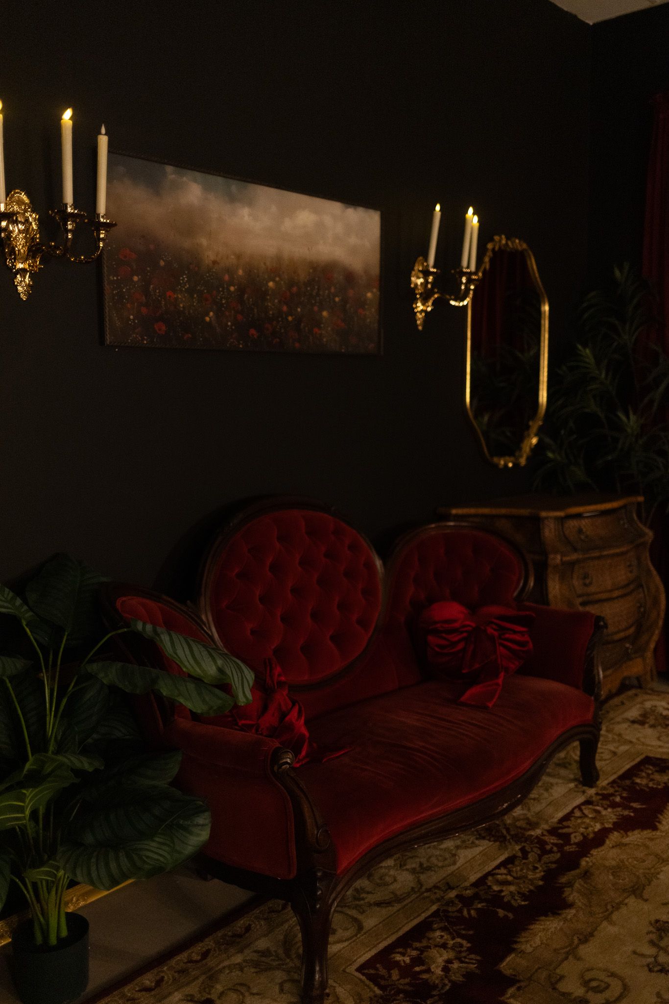 Elegant dark room with red velvet Victorian sofa, gold candelabras, floral painting, oval mirror, and lush green plants.