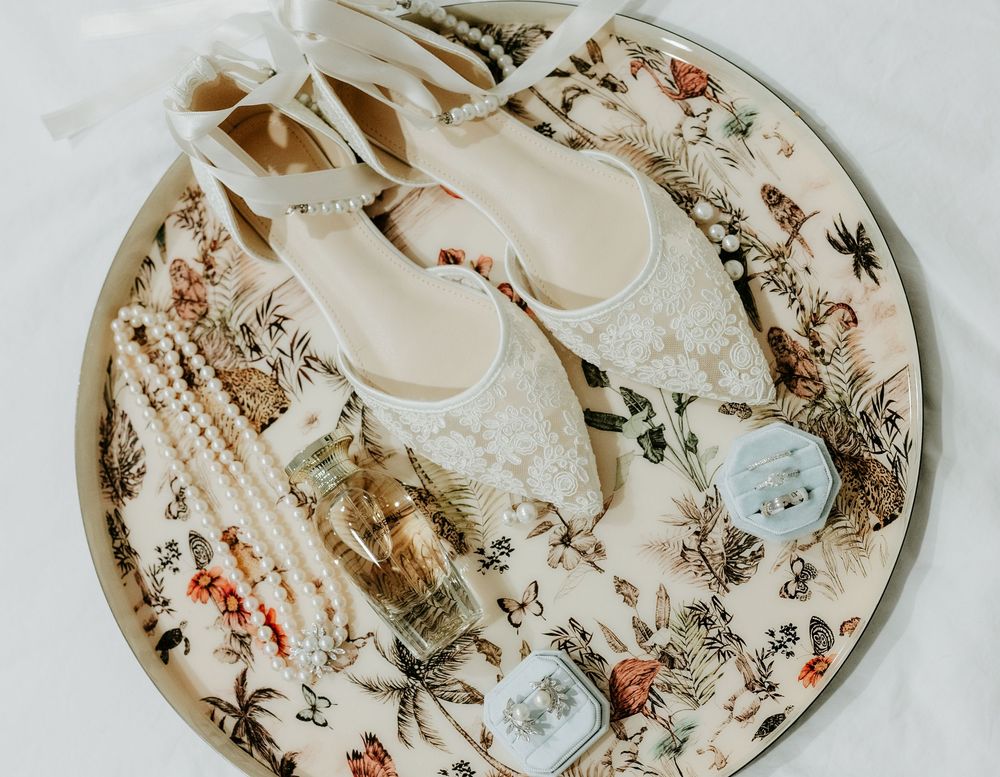Elegant bridal flat shoes with lace detail on floral tray with perfume, jewelry, and accessories.