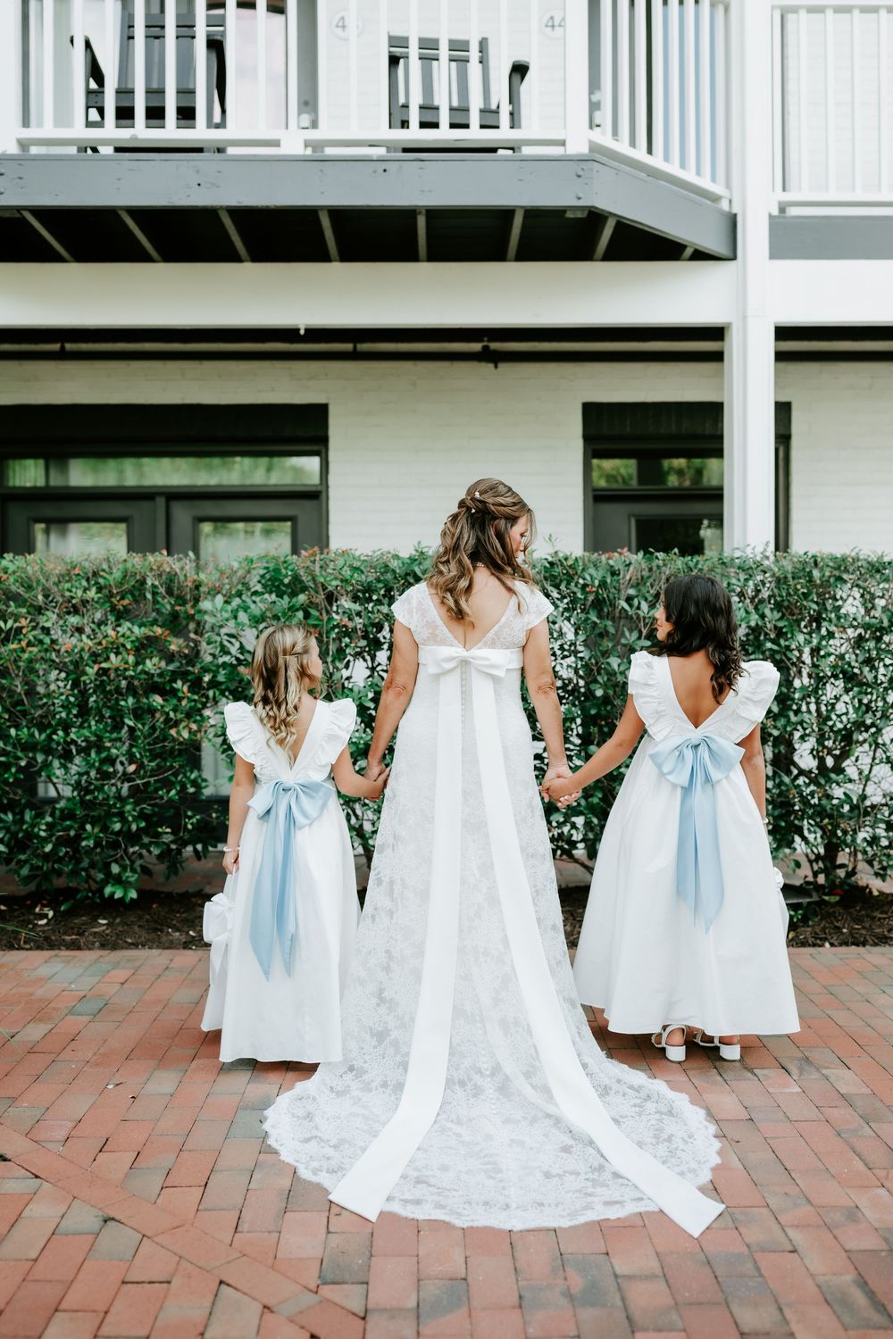 Bride in white gown with lace train holds hands with two flower girls in white dresses with blue bows outdoors.
