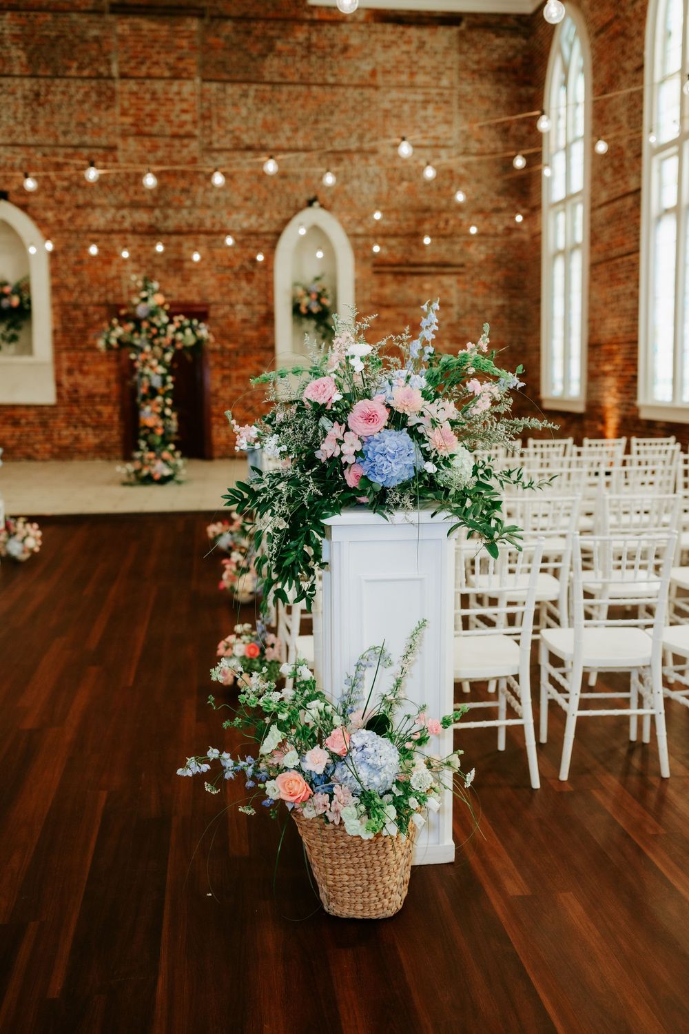 Elegant wedding ceremony hall with floral arrangements of pink and blue blooms on white pedestals and wicker baskets.