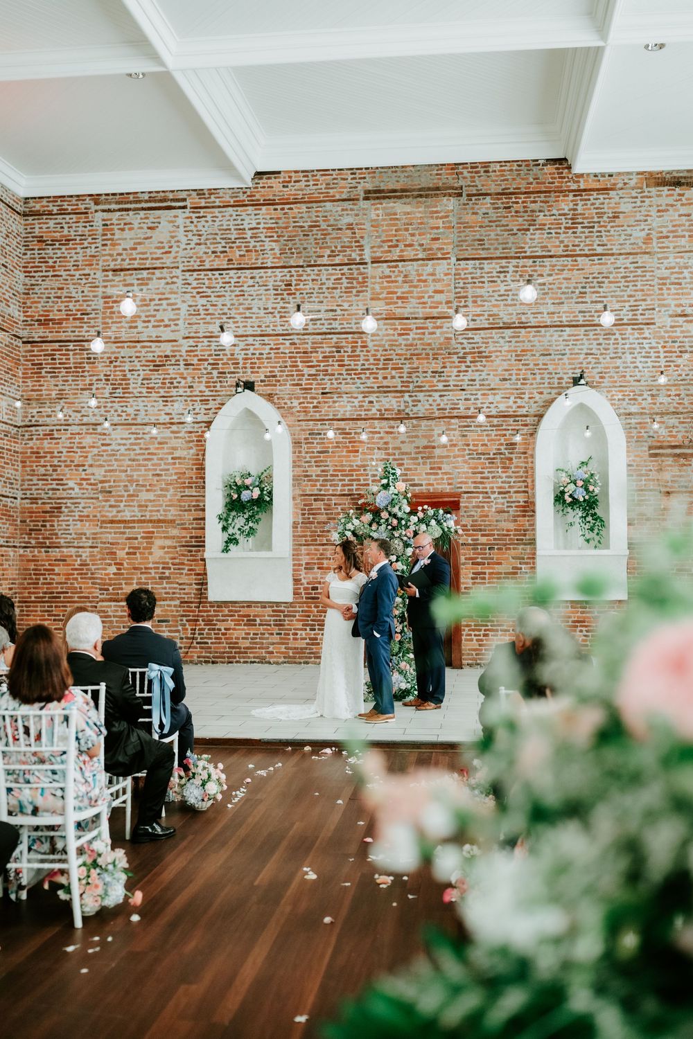 Couple exchanging vows at elegant indoor wedding ceremony with brick walls, floral arch, string lights, and seated guests.