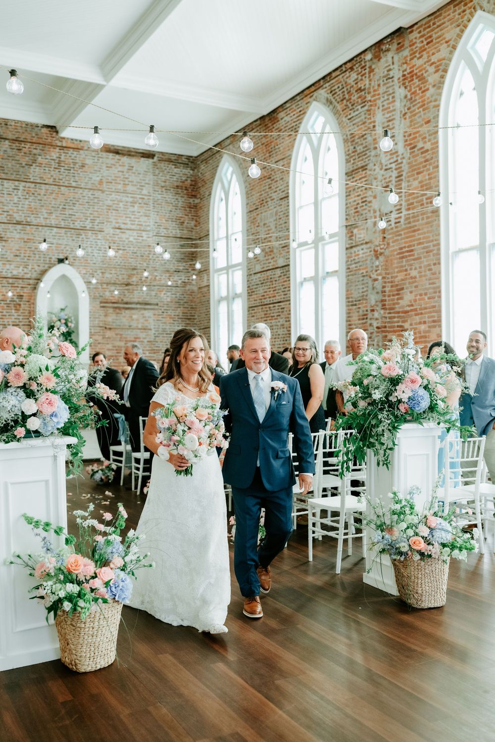 Bride walks down aisle with escort in elegant brick chapel adorned with pastel floral arrangements and string lights.