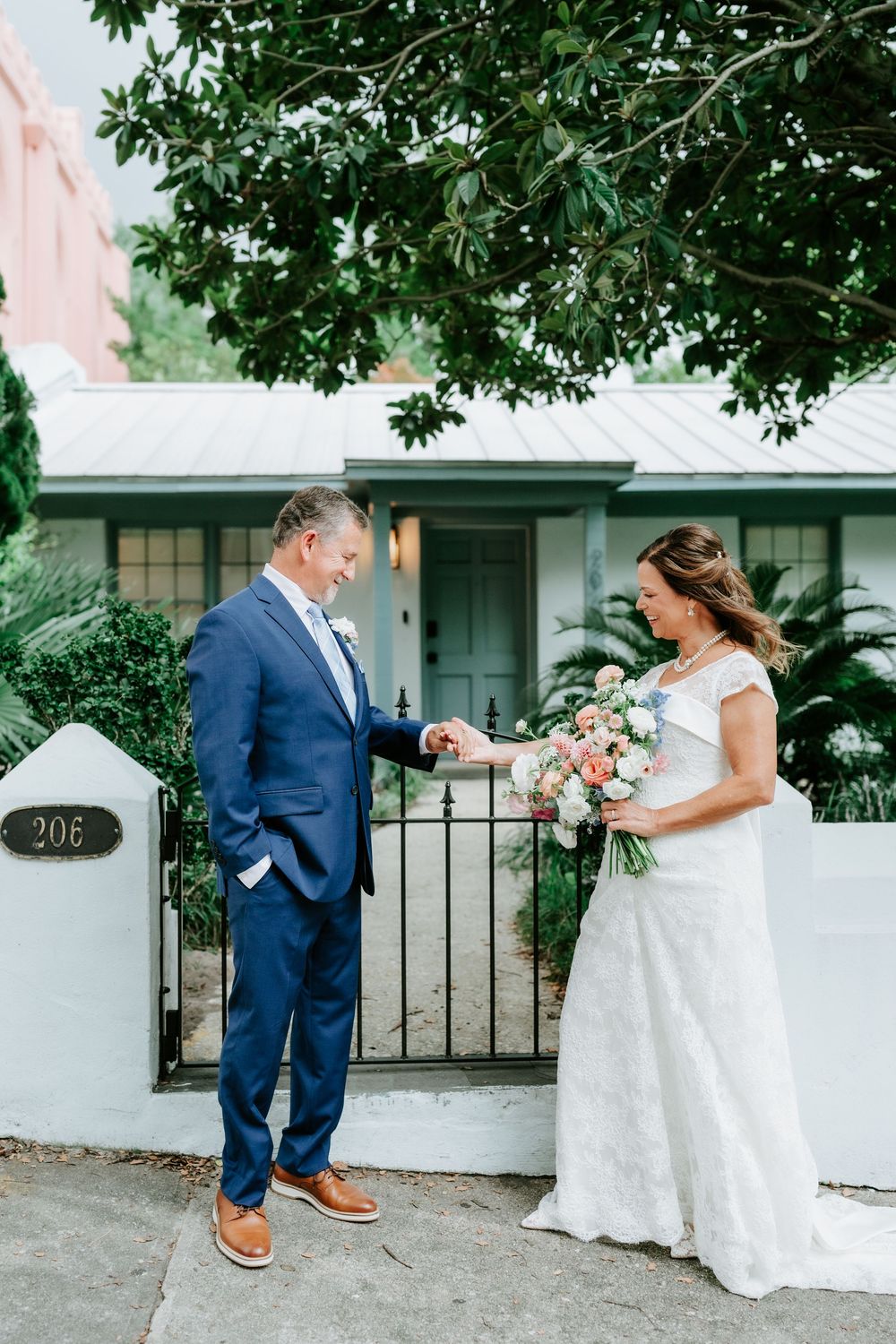 Bride and groom share a tender moment outside a charming home, she holds a pastel bouquet while he examines her ring.