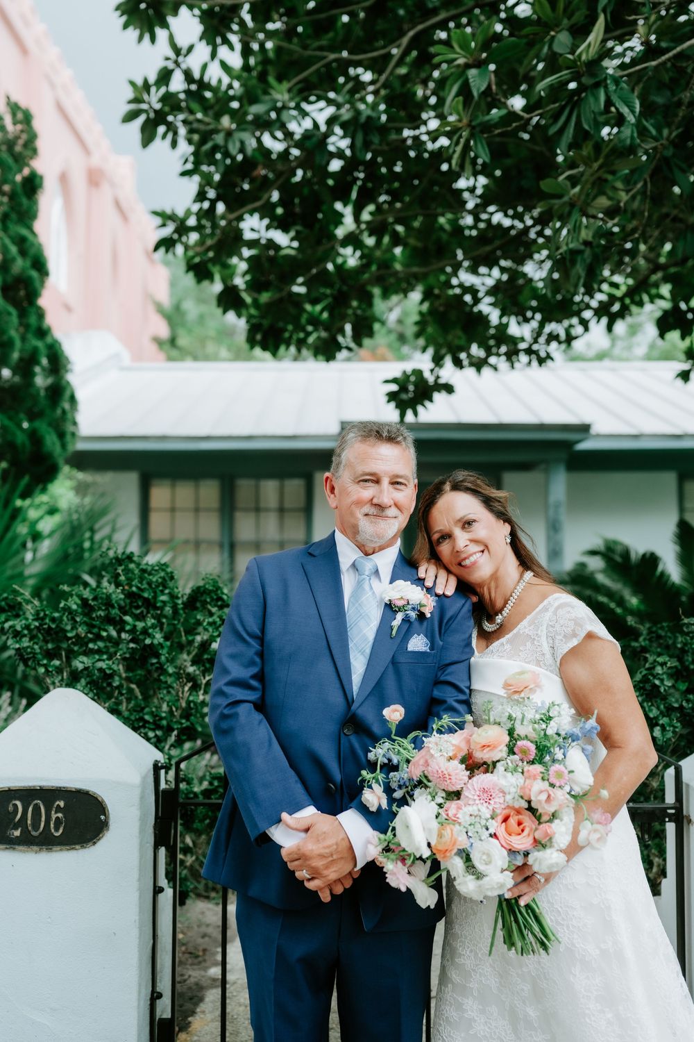 Bride and groom smiling together outdoors, she holds a pink and white bouquet, he wears a navy suit with boutonniere.