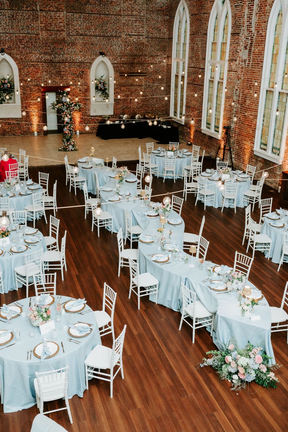 Elegant wedding reception in a brick venue with light blue tablecloths, white chiavari chairs, and floral centerpieces.