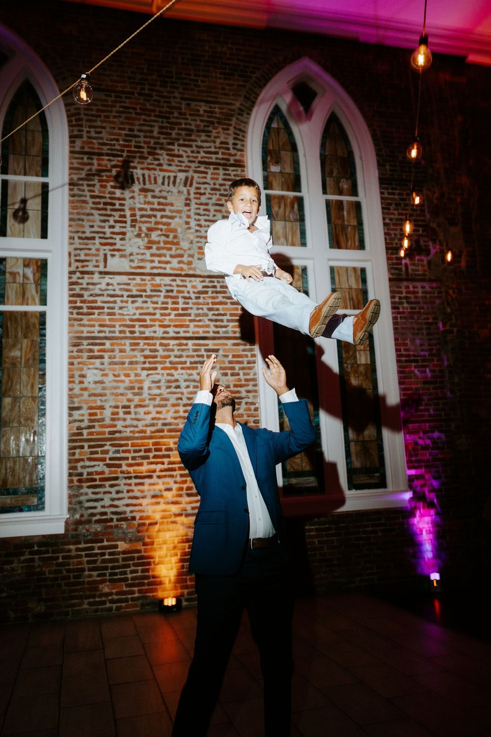 Man in suit tossing laughing boy in white suit into the air at elegant brick venue wedding reception.