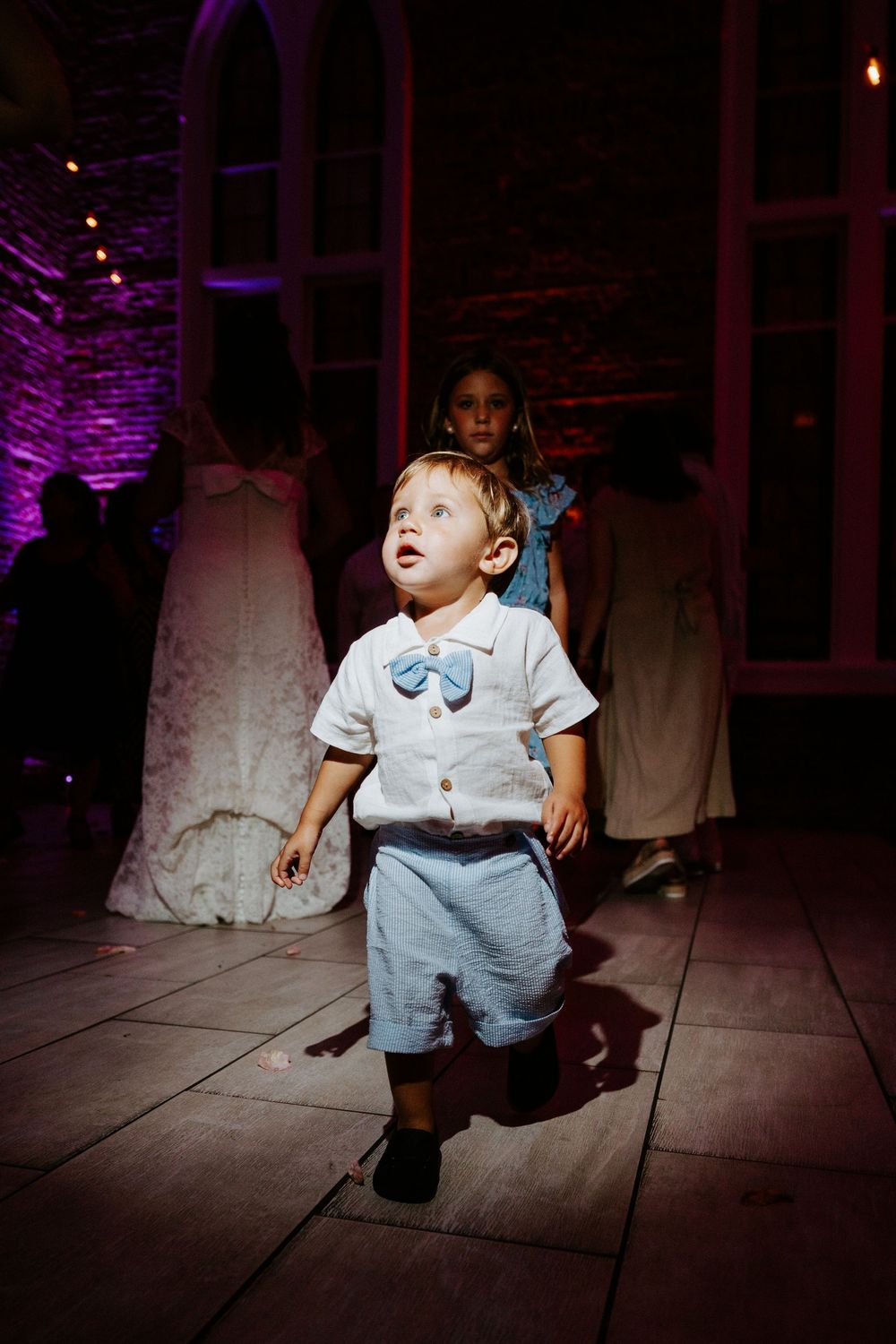Toddler boy in white shirt with blue bow tie and shorts walking on dance floor at wedding reception.