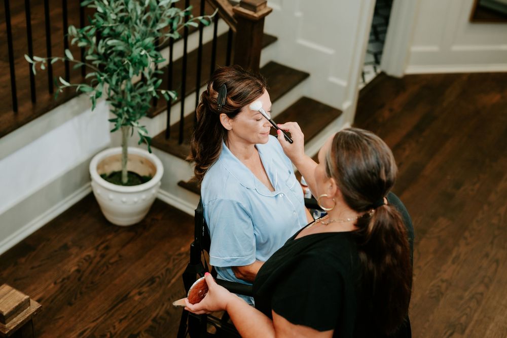 Makeup artist applying cosmetics to a woman in a blue robe near a staircase in an elegant home.