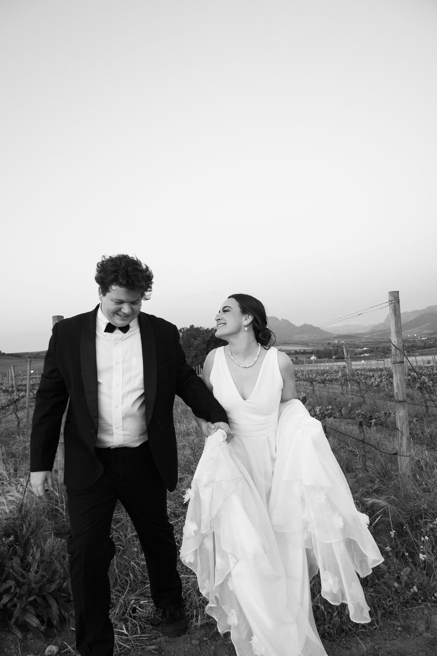 Black and white photo of a smiling bride in white gown and groom in tuxedo walking together in a vineyard.