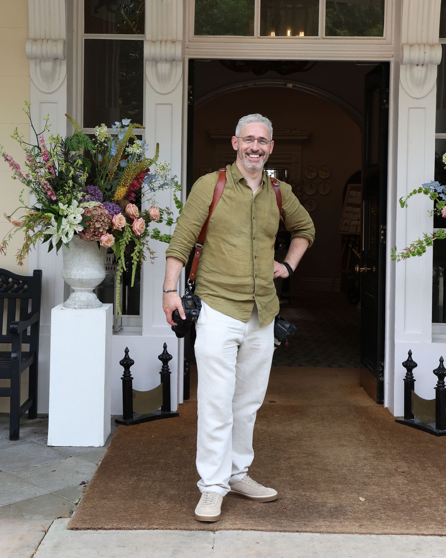 Smiling man in olive shirt and white pants stands at elegant building entrance with floral arrangement.