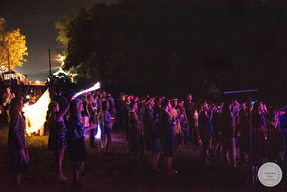 Crowd of people dancing outdoors at night festival with colorful lights and bonfire in background.