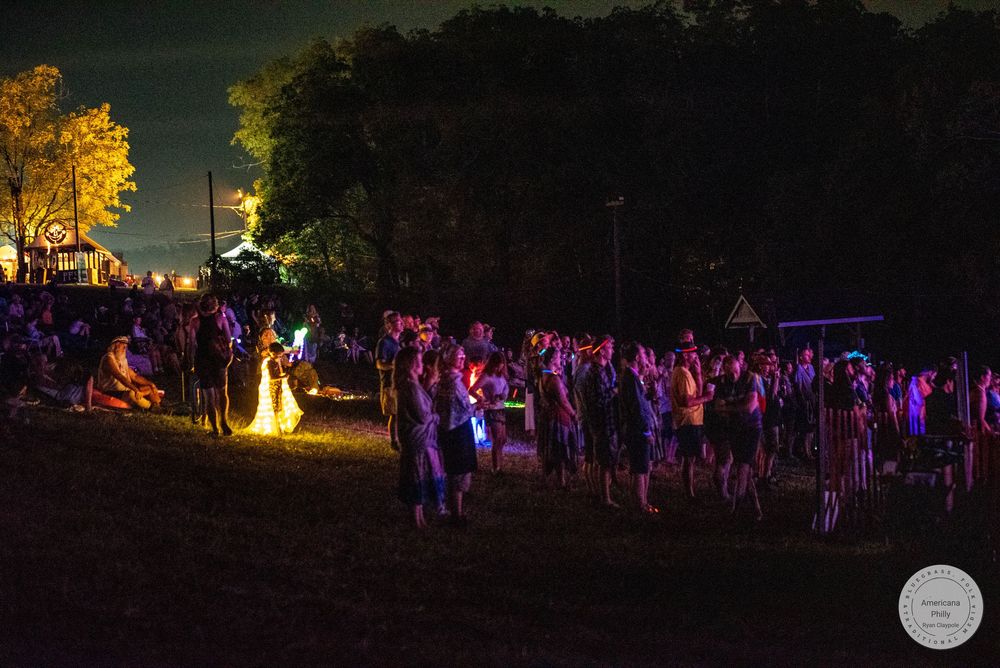Crowd of festival-goers gathered at night outdoors with colorful lights and a bonfire glowing in the background.