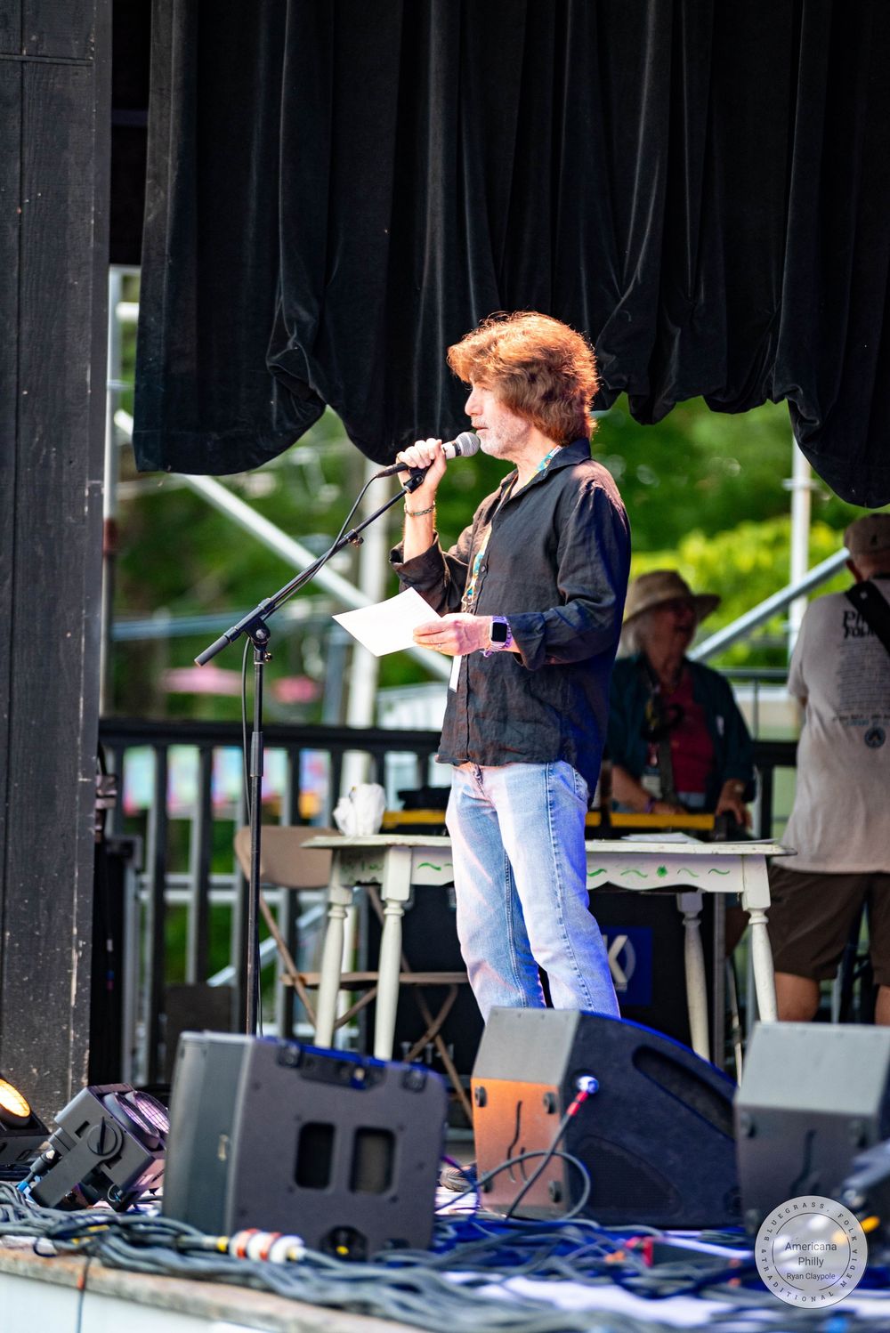 Woman with curly red hair speaks into microphone on outdoor stage, holding papers, wearing black jacket and jeans.