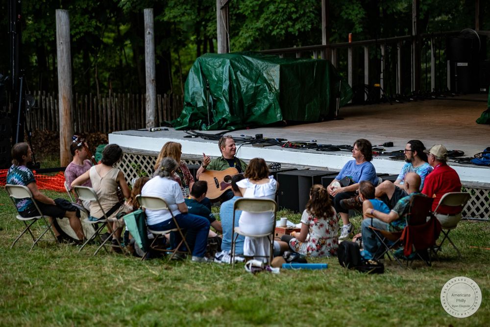 Group of people sitting in a circle on grass playing guitar at an outdoor music gathering near a stage.