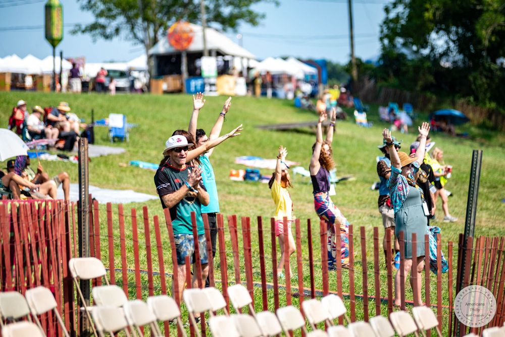 Festival crowd cheering with raised hands on sunny green lawn near white tents and wooden fence at outdoor music event.