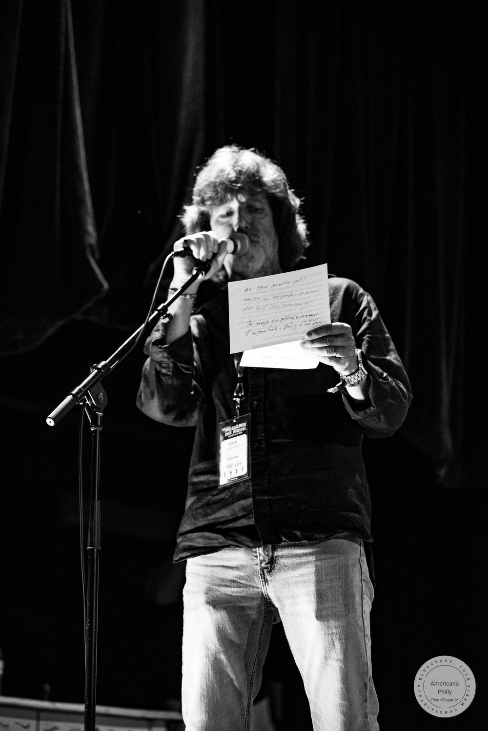 Black and white photo of a man speaking at a microphone on stage, holding a handwritten note, wearing a lanyard.