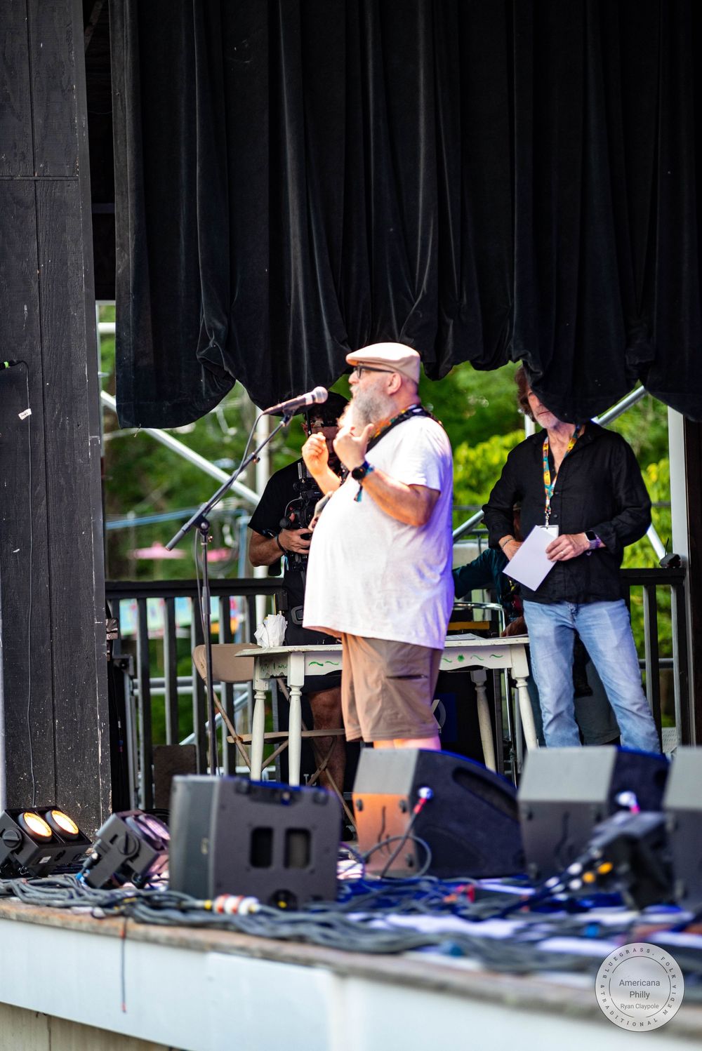 Bearded man in white shirt speaks at outdoor stage microphone during live music event with band members behind him.