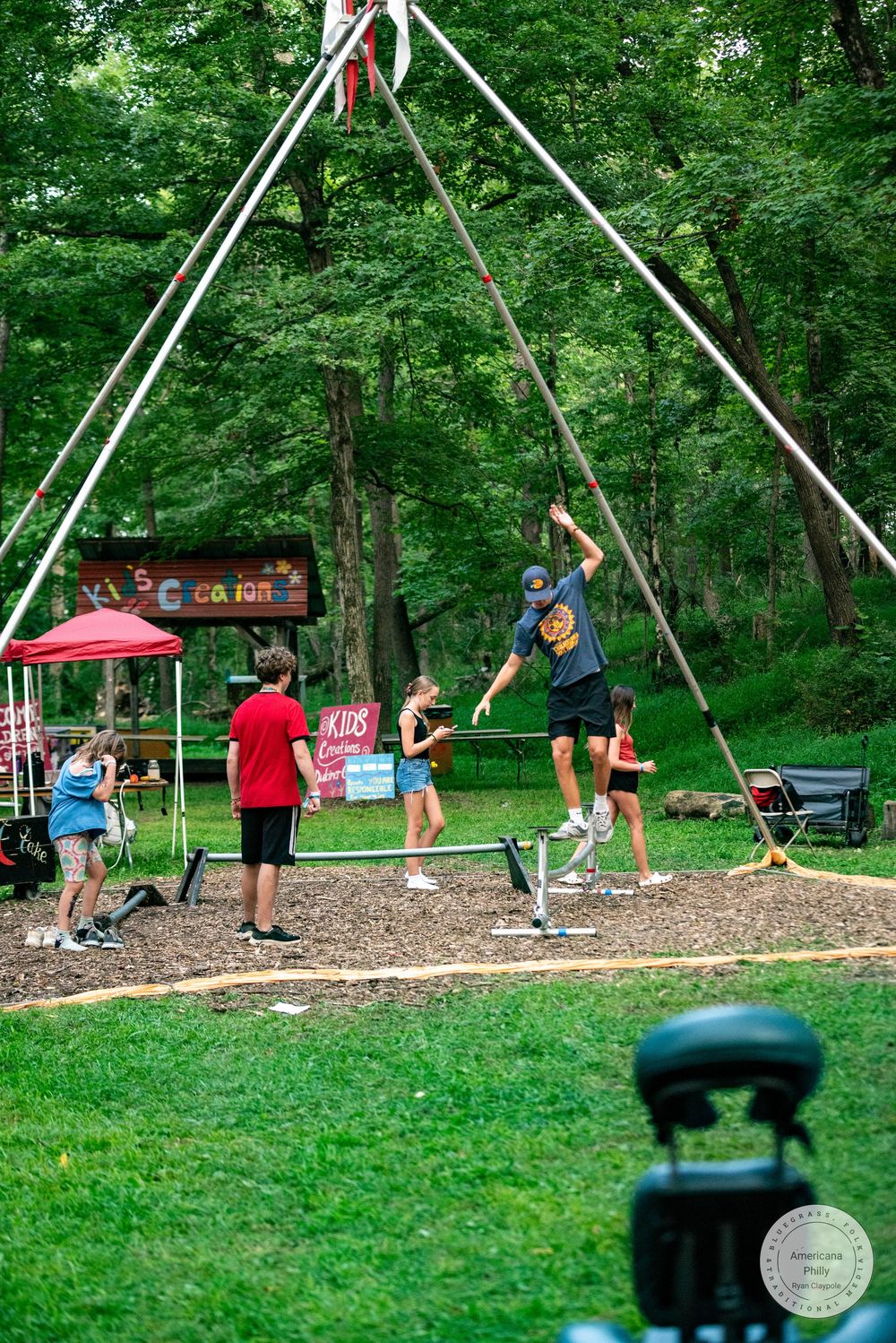People enjoy outdoor activities at a summer camp park with a bungee trampoline structure surrounded by lush green trees.