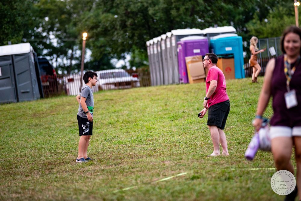 Two people playing catch on a grassy hill at an outdoor festival with portable toilets in the background.