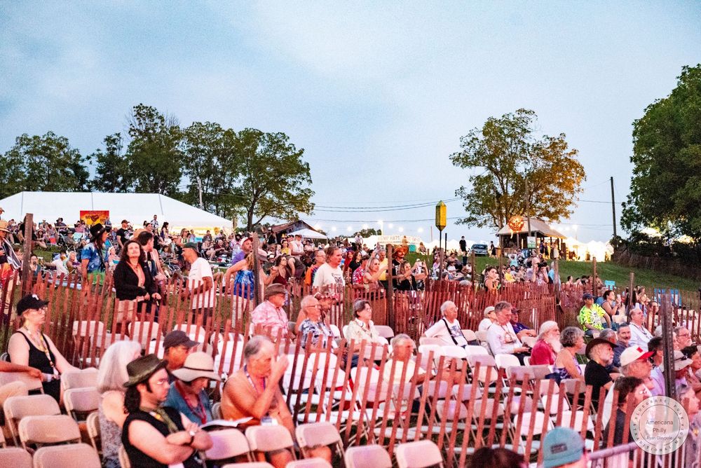 Large outdoor amphitheater crowd enjoying a live music festival event at dusk with trees and white tents in background.