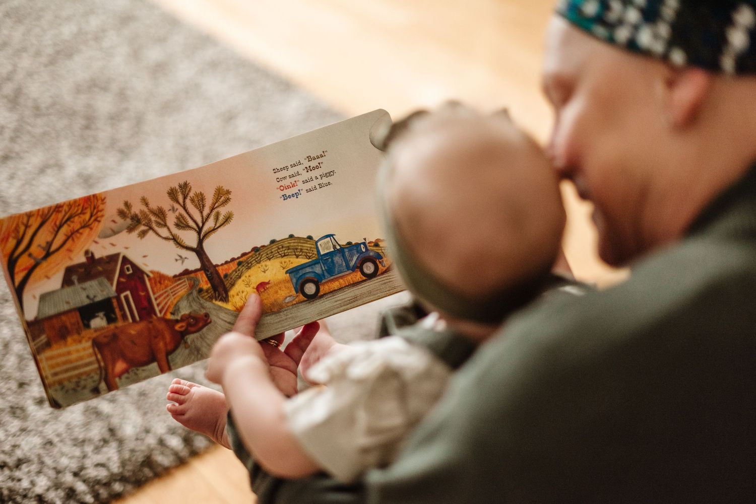 Adult reading a colorful farm-themed picture book to an infant while sitting on a floor.