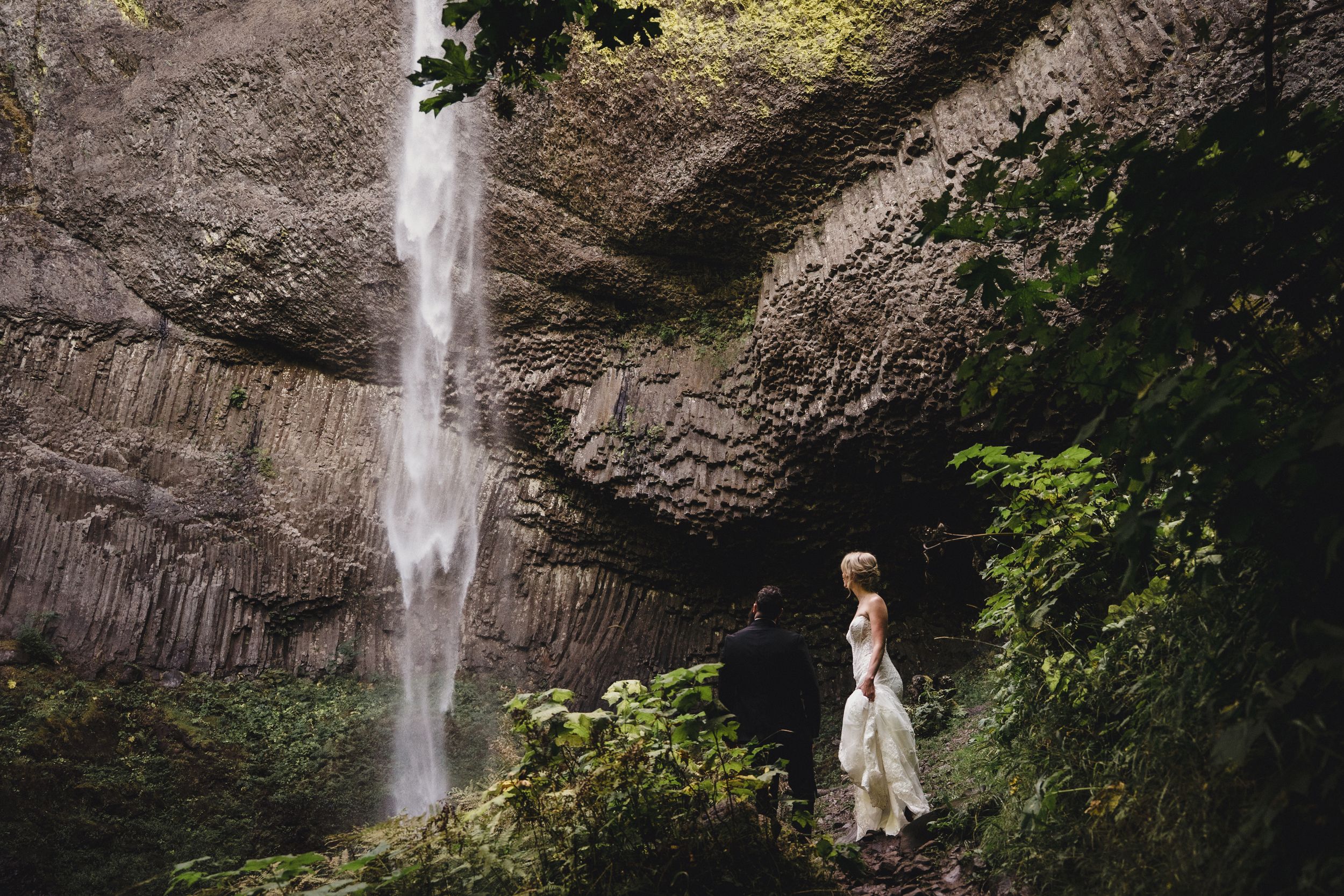Bride in white dress standing before a majestic waterfall cascading down rocky cliffs surrounded by lush greenery.