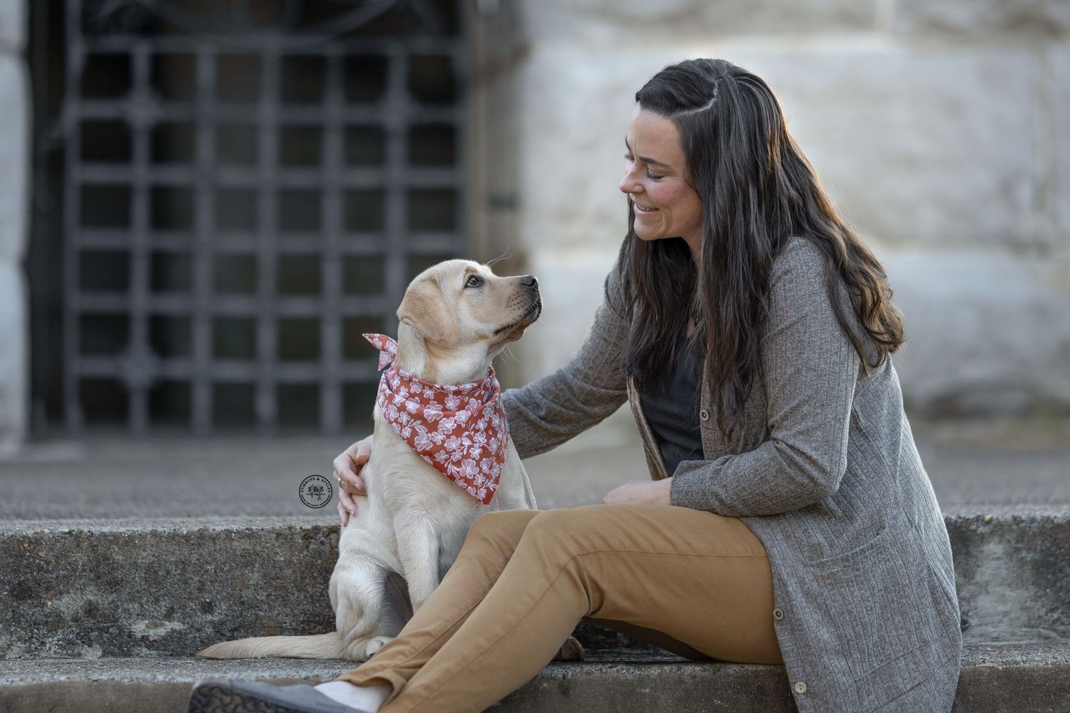 A smiling woman with long dark hair pets a yellow Labrador puppy wearing a red bandana while sitting outdoors.