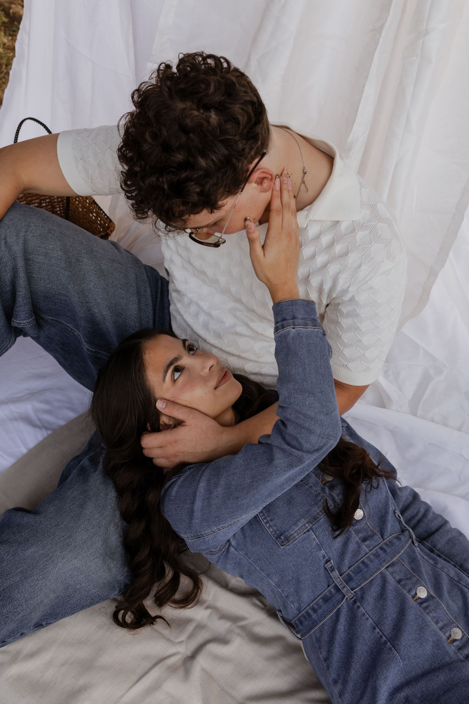 A couple shares a tender moment, with one person lying down while the other leans in closely on a white fabric backdrop.
