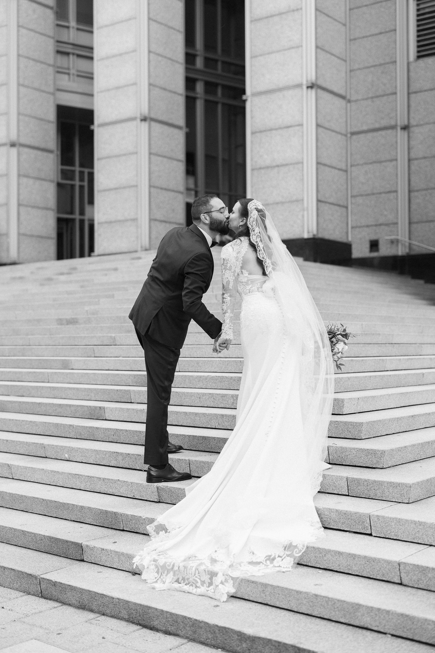 Black and white photo of bride and groom sharing a kiss on grand stone steps, bride in lace gown with veil.