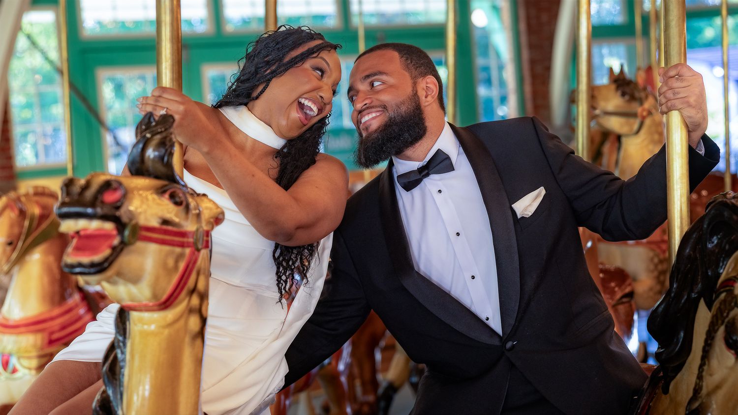 A joyful couple in wedding attire laughing together on a colorful carousel, groom in tuxedo and bride in white gown.