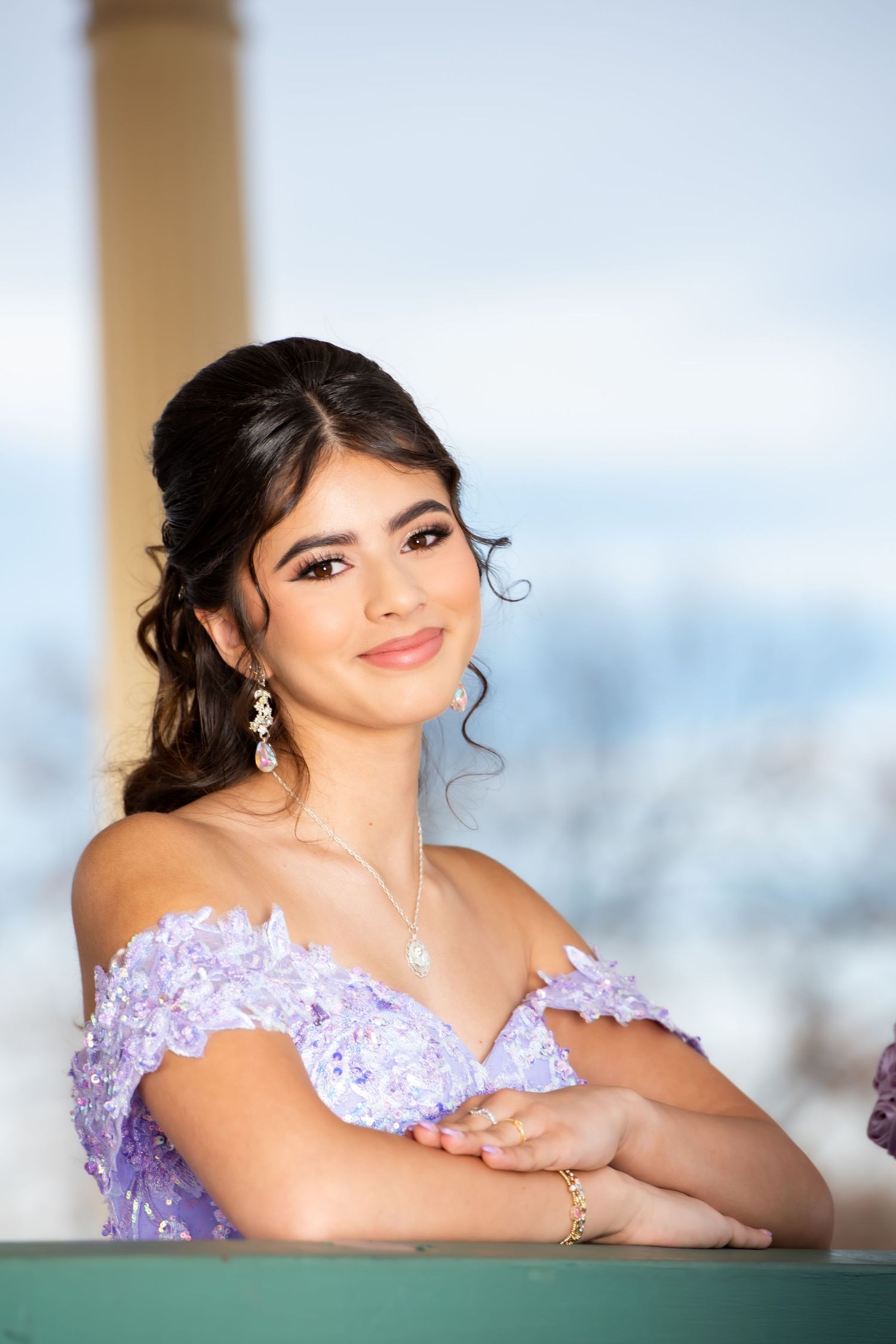 Young woman in lavender off-shoulder quinceañera gown with floral lace smiling elegantly at camera.