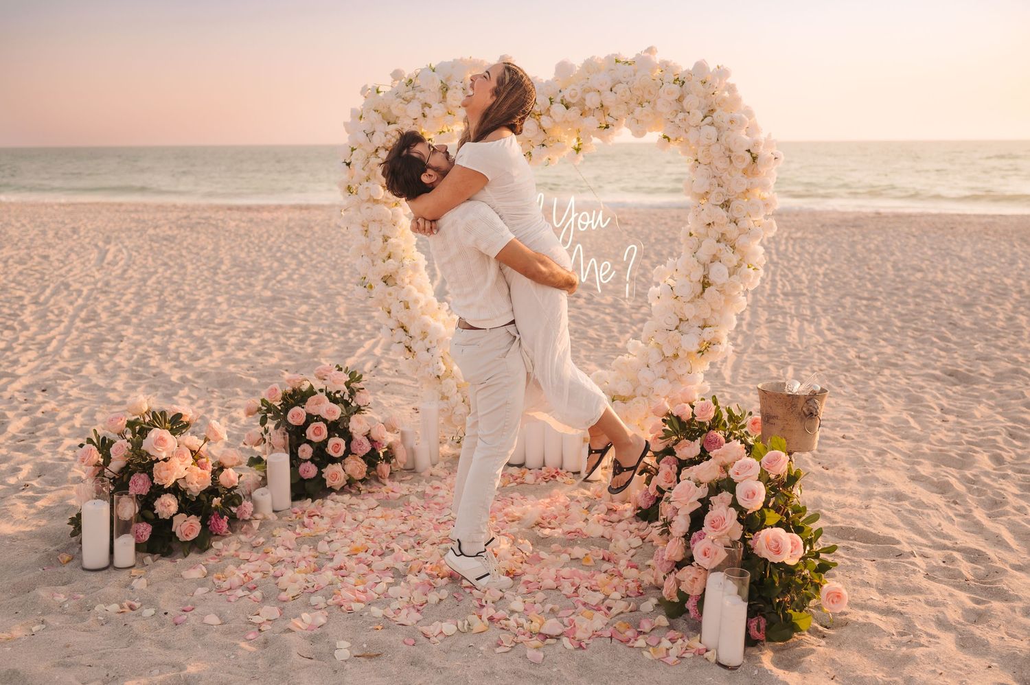 A joyful couple celebrates a beach marriage proposal surrounded by a floral heart arch and roses.