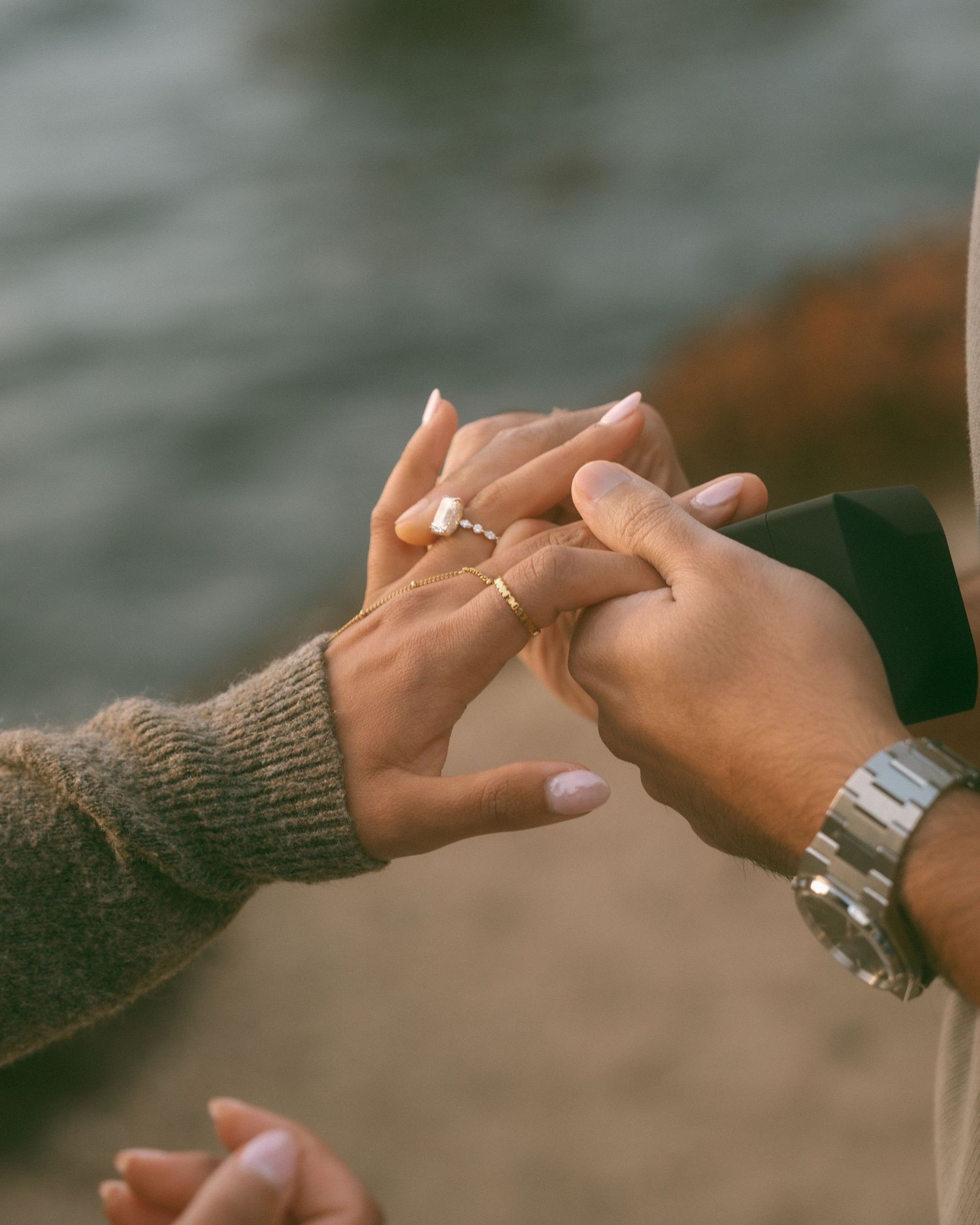 A person placing a diamond engagement ring on their partner's finger outdoors near water.