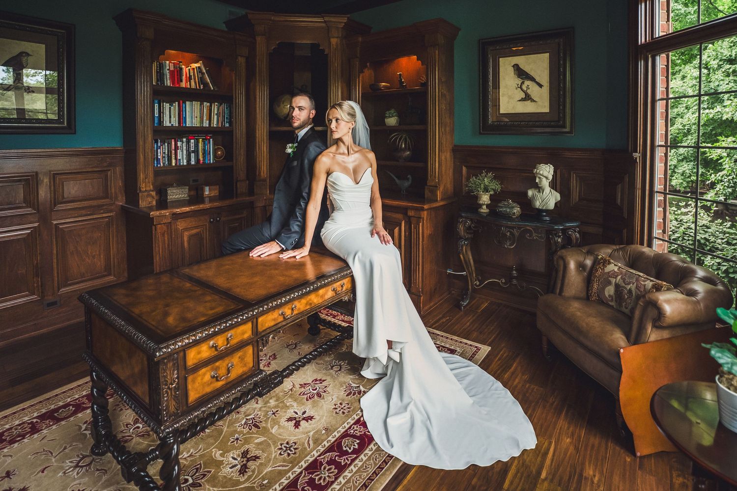 Bride in a flowing white gown sits on an ornate wooden desk beside her groom in a classic study with green walls and leather armchairs.