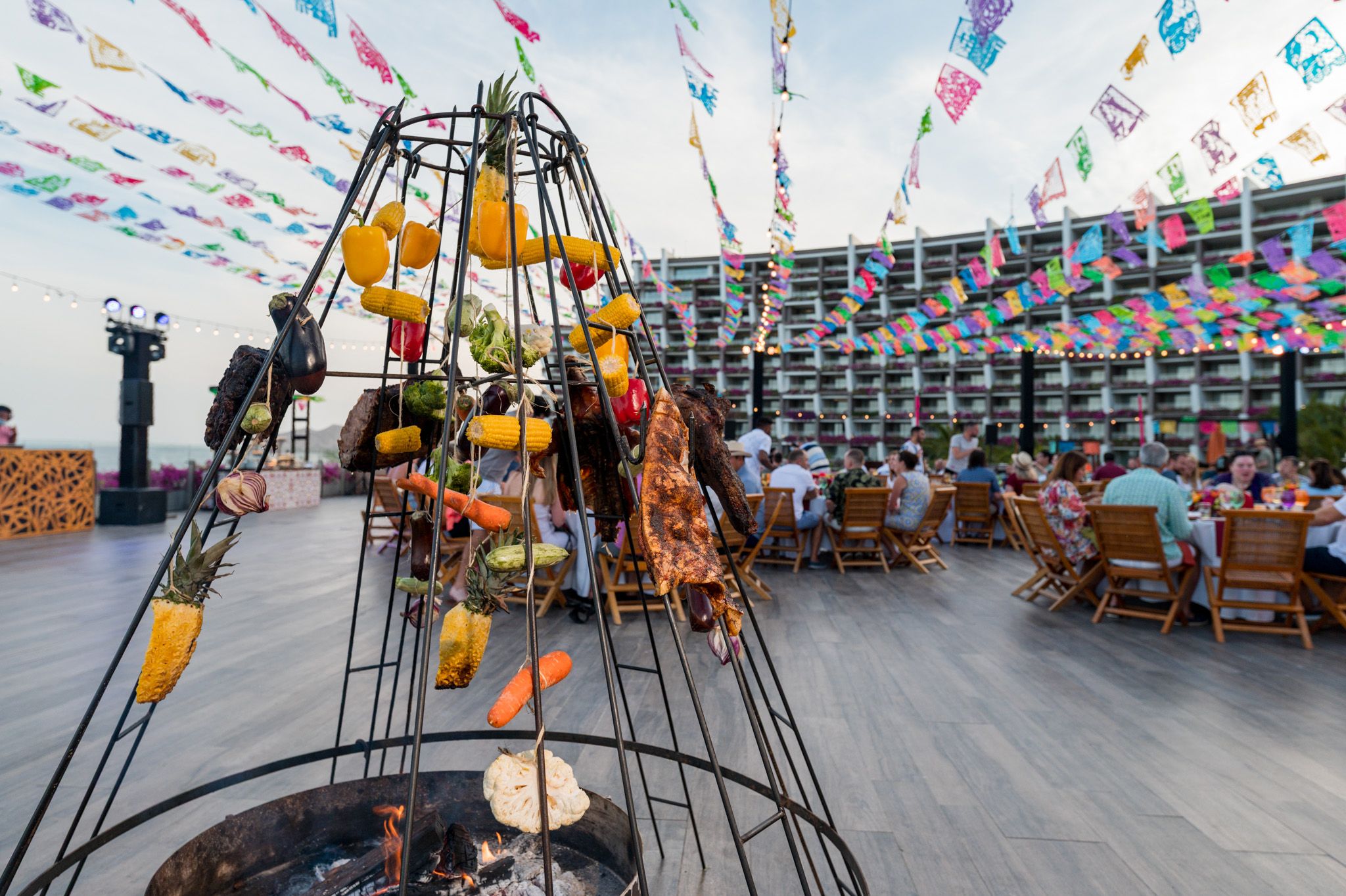 Outdoor BBQ grill with meats and peppers over fire pit, colorful bunting flags, hotel terrace dining at dusk.