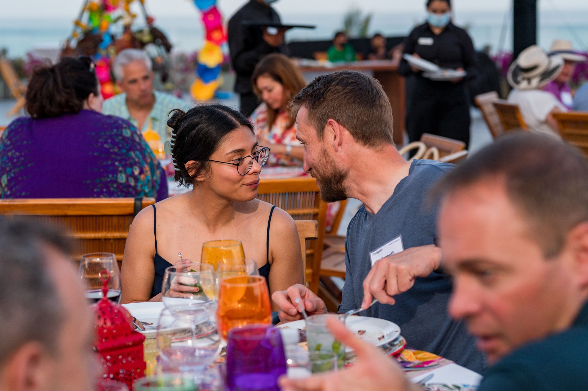 Couple sharing a romantic moment at an outdoor beachside dinner party with colorful drinks and decorations.