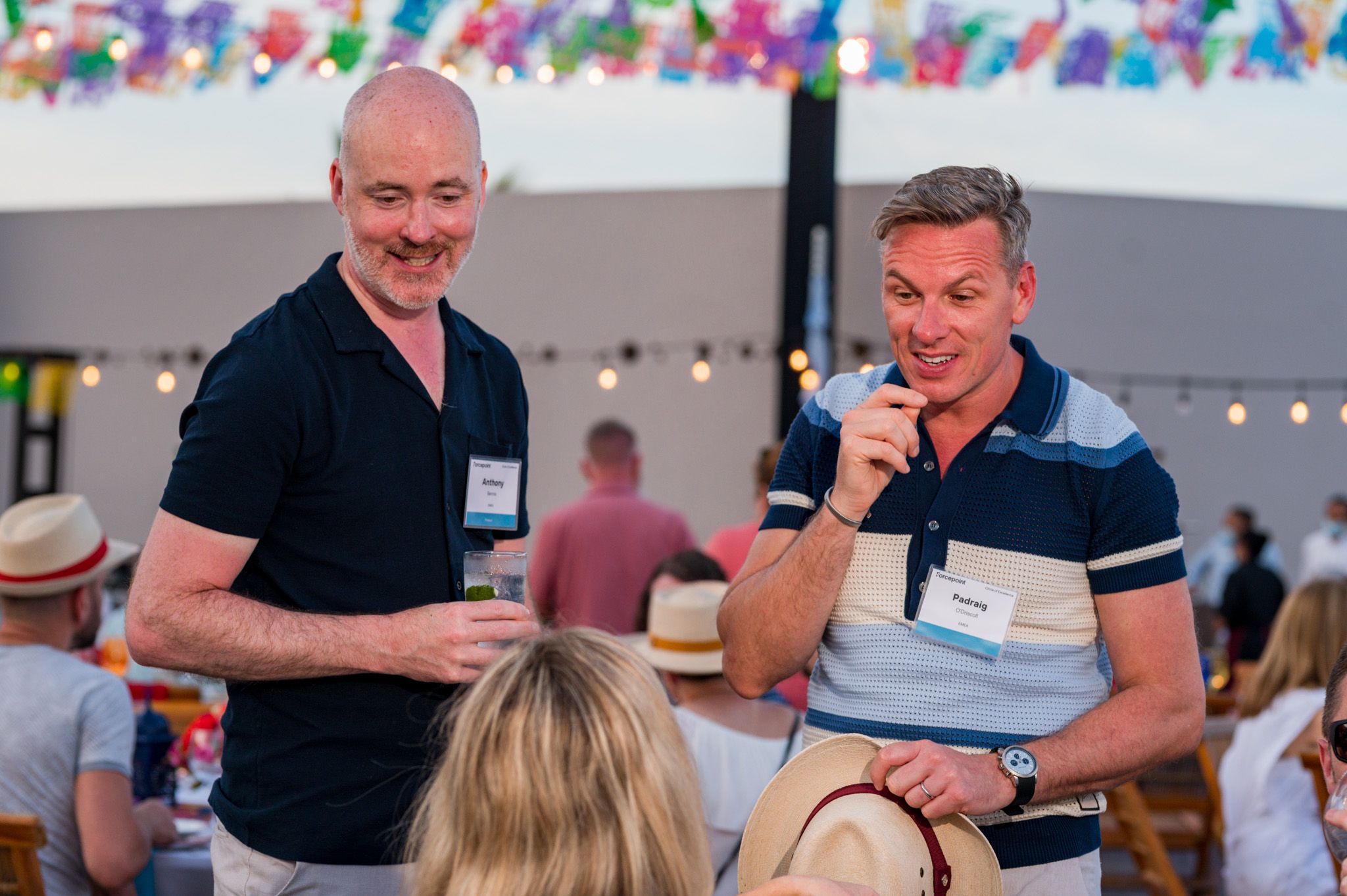 Two men smiling and chatting at an outdoor social event, one holding a drink, with festive decorations in the background.